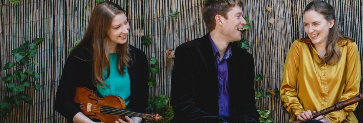 Photograph of the Ensemble Hesperi group. On the left is Magdalena Loth-Hill who is dressed in blue with long brown hair and holding her violin; next to her also dressed in blue is Thomas Allery with short brown hair, who will play the piano and next to him is Mary Jannet-Leith, who has long blond hair and is dressed in yellow and holding a recorder