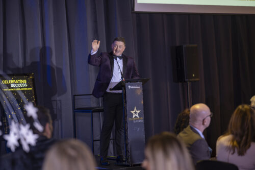 a man wearing a dinner jacket stands on a podium, his arm is raised in the air
