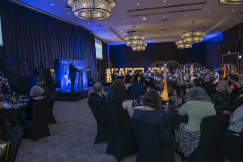 a large room with people seated around tables. a man is standing on the stage speaking into a microphone