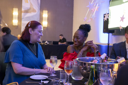 two people seated at a table talk to one another whilst smiling