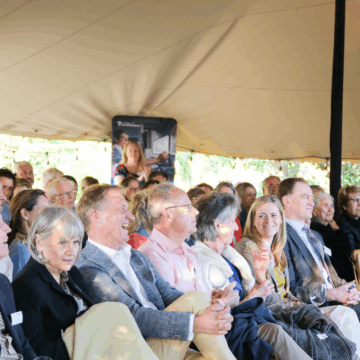 A group of smartly dressed people, sat on wooden chairs under a beige canopy.
