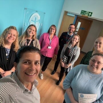 a selfie photograph of a group of people standing in a corridor. on the wall is a perspex logo with the words "lifelab".