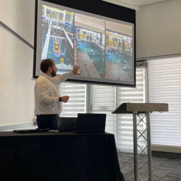 a man stands at a lectern and gestures to the presentation on the screen behind him, with images of a tank of water and model boats on it.