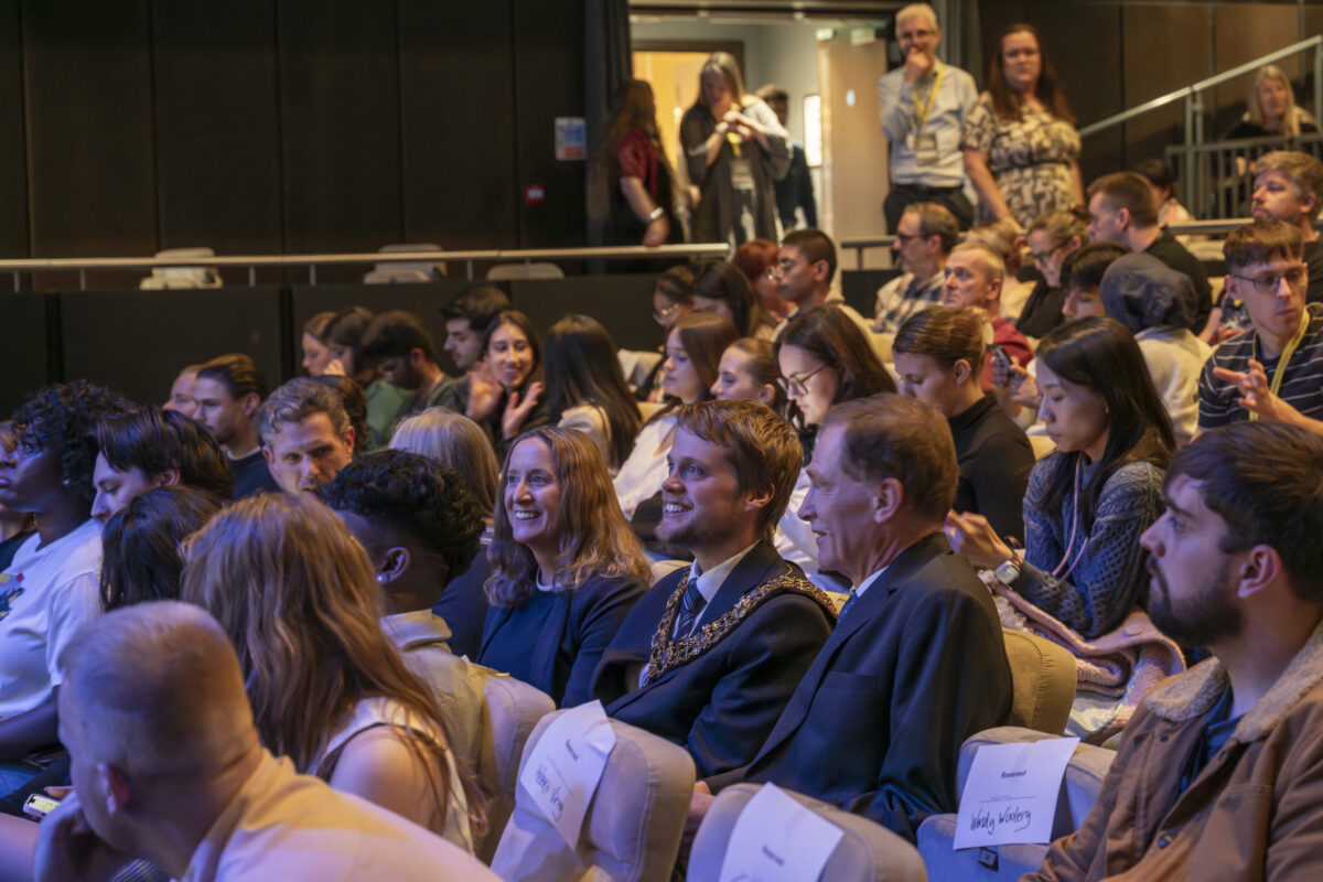 A Mayor, wearing a suit and a civic chain, is sat smiling in a busy audience,
