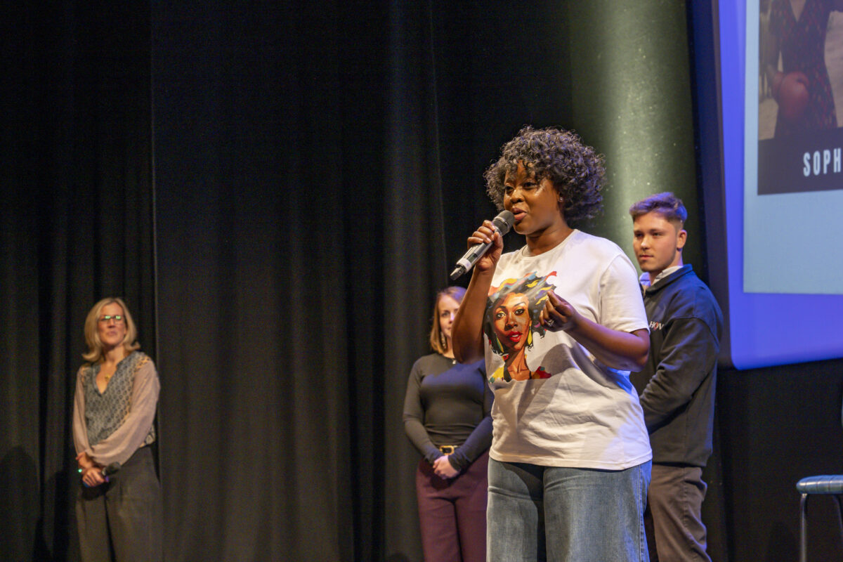 A woman with short, dark curly hair is speaking into a microphone on stage.