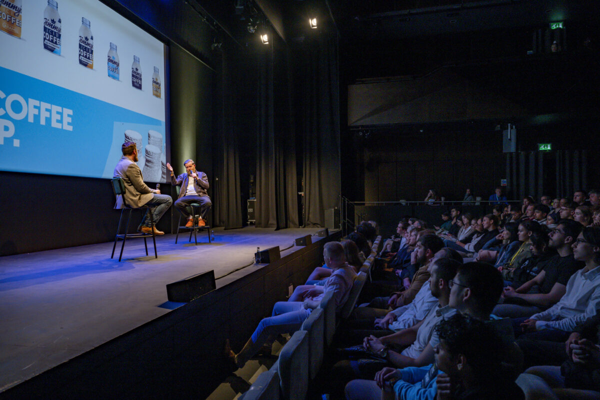 A large audience is watching two men, sat on stools, speak on stage.