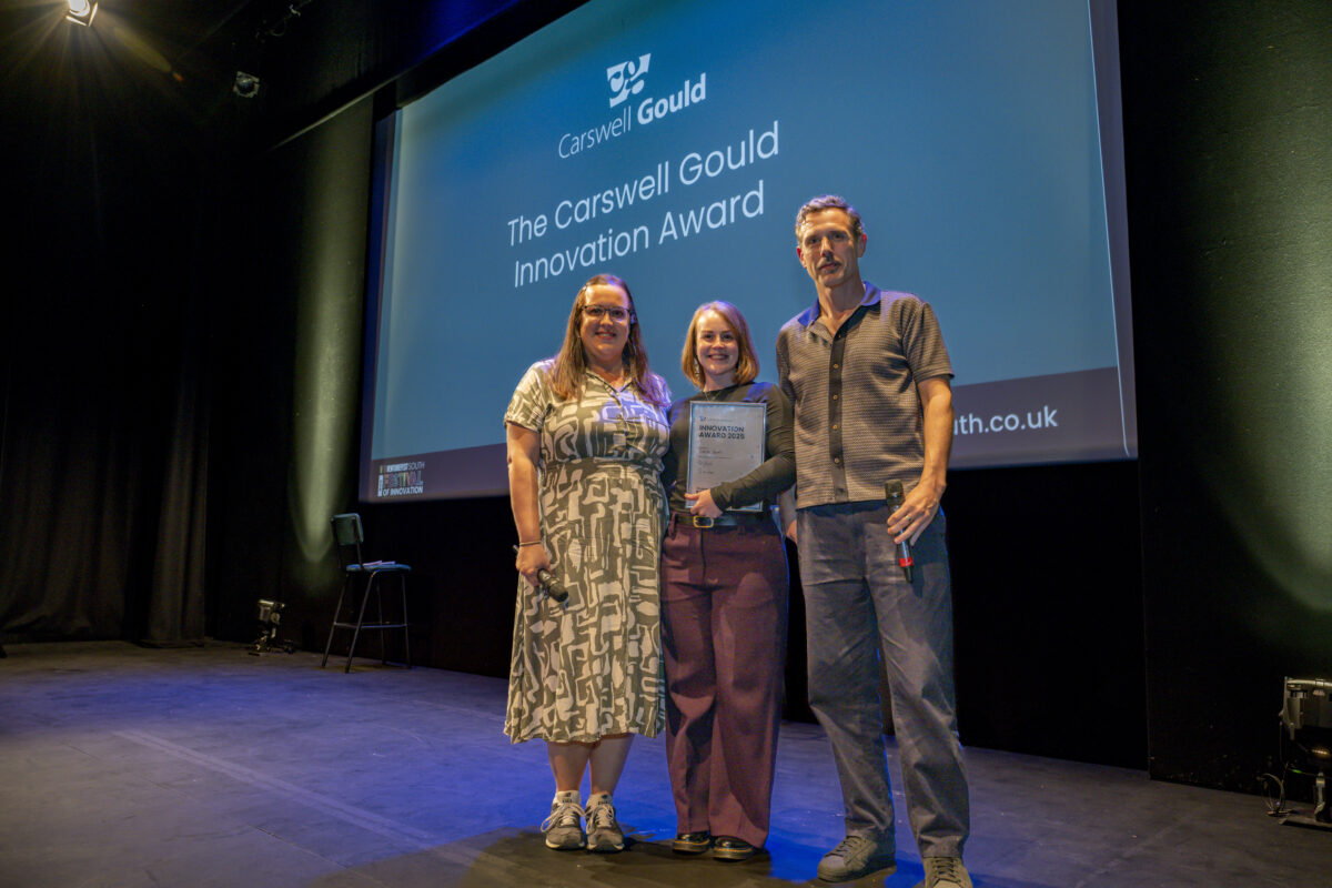 Three people are stood on stage - the woman in the middle is smiling and holding an award.