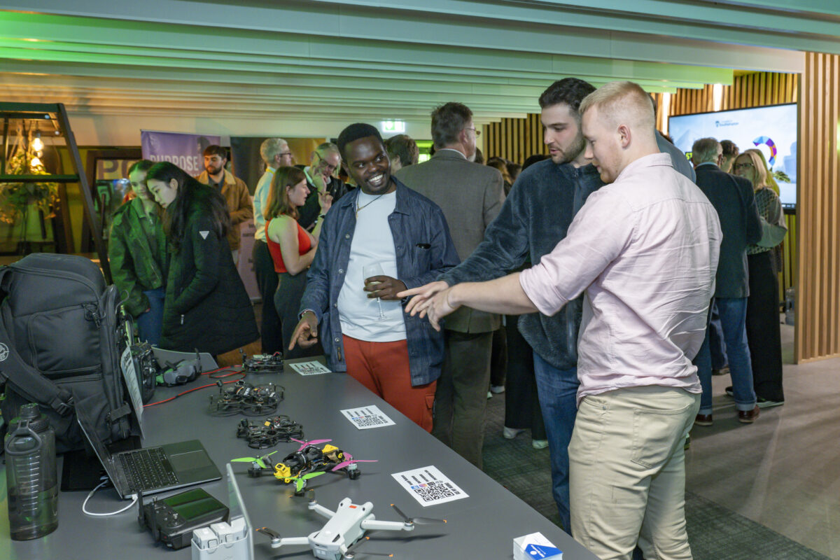 Three men are admiring a few different drones which are laid out on a table.