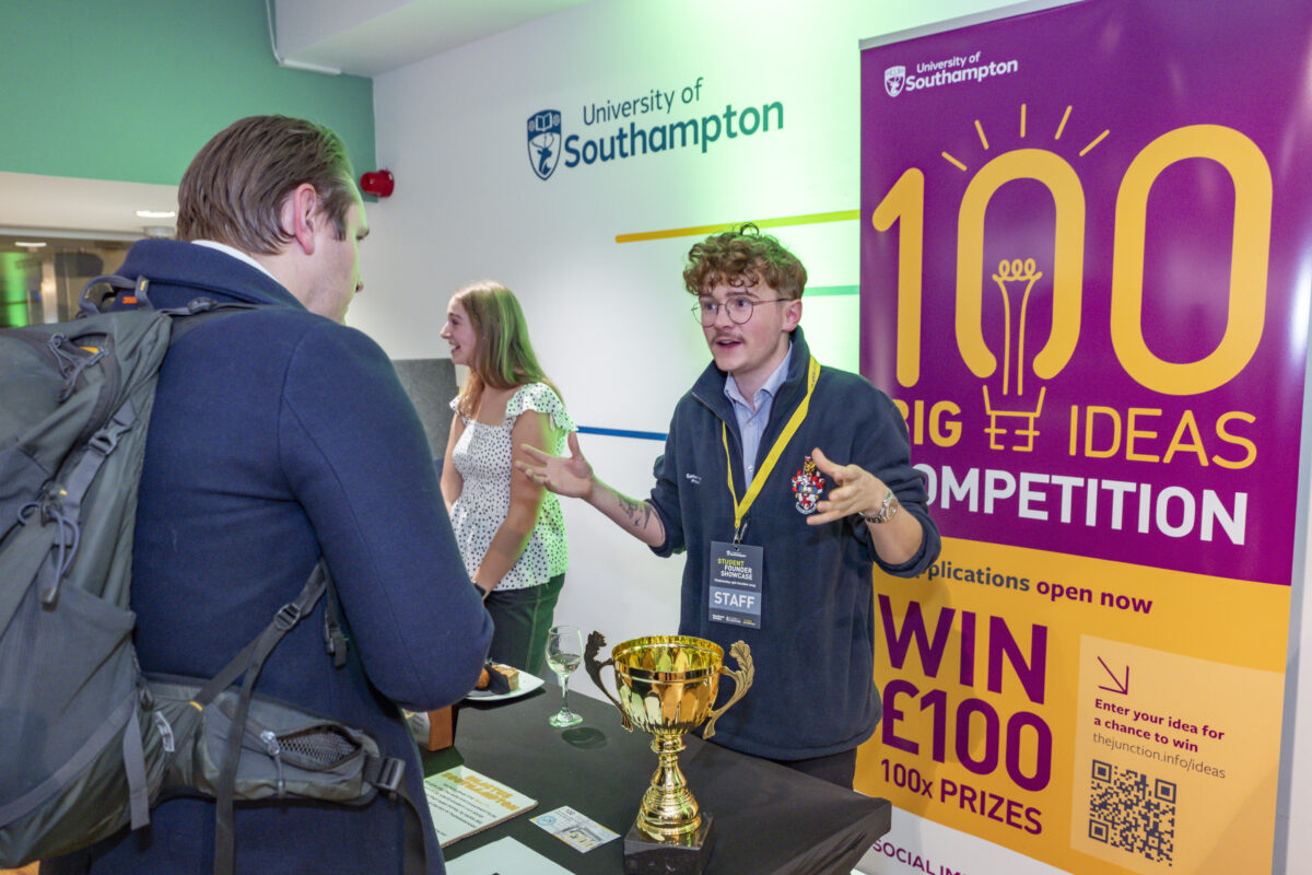A curly-haired man is stood behind a table with a gold trophy on. He is speaking to another man who is stood on the other side of the table.