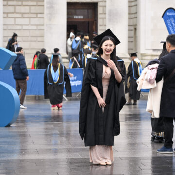 A woman in a graduation cap and gown is posing for a photo, laughing with her hand raised towards her face.