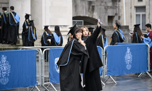 Two friends wearing cap and gowns, taking a photo though the back camera of their phone.