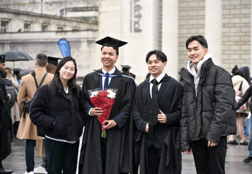 A group of young people stood together, two are wearing Graduation gowns and one is holding a bouquet of flowers.