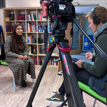 A woman wearing a hijab is sat in front of a camera, a man is also sat in front of her, looking at his phone.