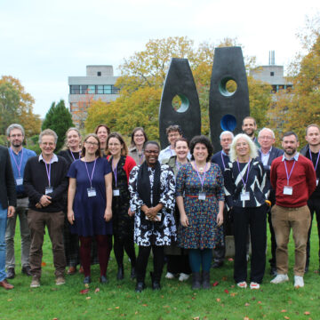 Three rows of people and stood smiling outside, in front of a tall sculpture with holes in.