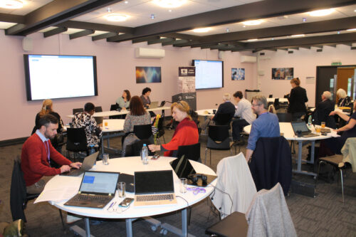 A room with people sat at white, round tables - two projectors are at the front of the room.