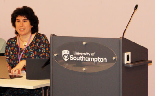 A lady with short, black hair is sat next to a grey podium with the University of Southampton Logo on.