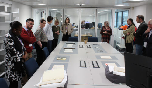 A group of people are stood round a grey, rectangular table, looking at paper laid out on it.
