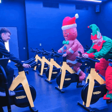 A spin class studio with an instructor at the front, facing the class which has someone dressed as an elf, next to a gingerbread man in.