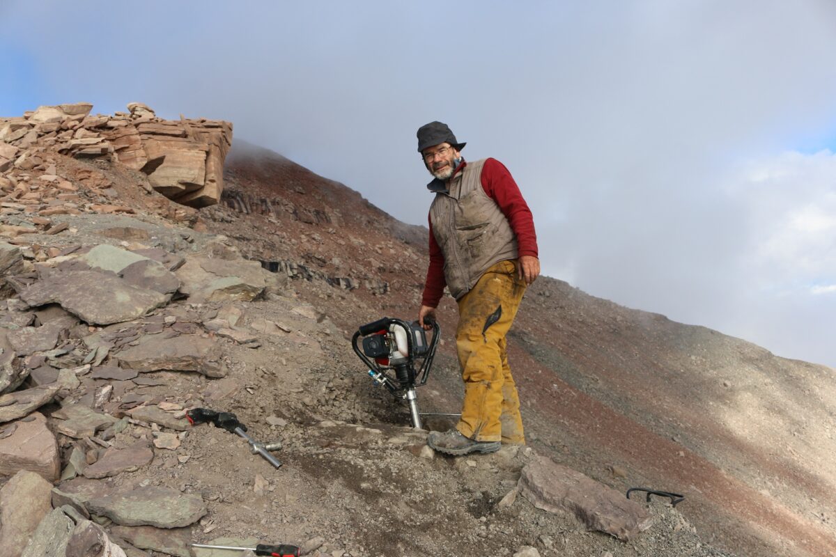 A man with dirty clothes stood on rocky, mountainous terrain, holding a drill in the ground.