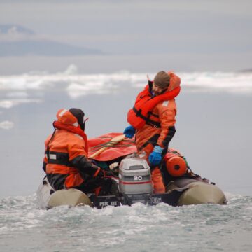 Two men wearing orange life jackers in a small boat in a large body of water.