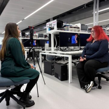 Two woman are sat opposite each other in a lab, the woman on the right has pink hair and is being interviewed.