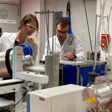 Two people wearing lab coats - the woman on the left is holding a cannister and pouring it in a container.