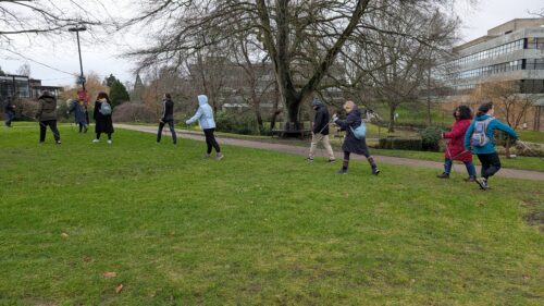 9 people with walking poles are walking around an area of grass on campus