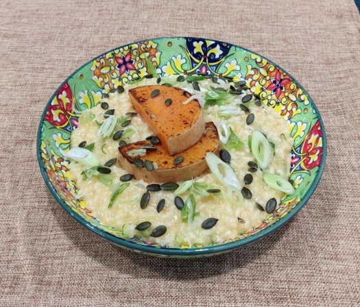 A colourful patterned bowl with creamy risotto, pumpkin seeds, chopped spring onions and 2 slices of roasted pumpkin on top