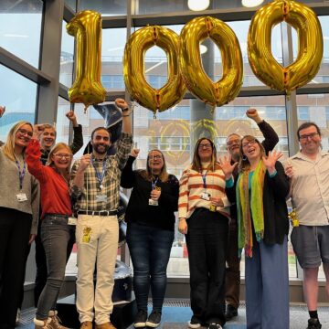 A group of people are stood in a line with their hands up, four of them are holding the strings for four balloons which make up the number '1000'.