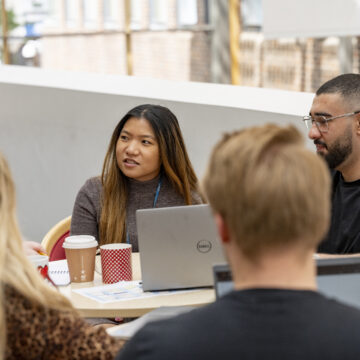A group of 5 people sit around a table, one of them is speaking and the others are looking towards them listening. On the table in front of them are number of laptops and coffee cups.