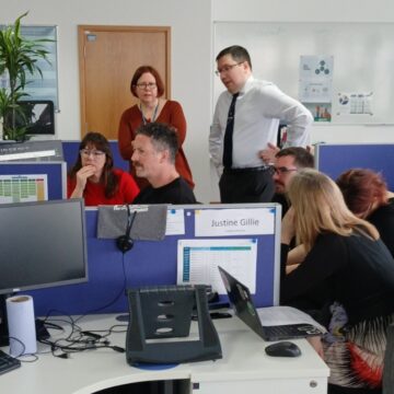 A group of seven people are huddled round a desk in an open office - two people are stood and the rest are sat.