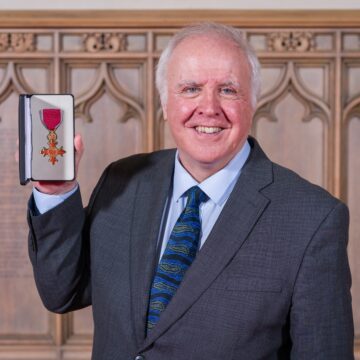 A man wearing a suit is smiling widely whilst holding up a medal, displayed in a small box.