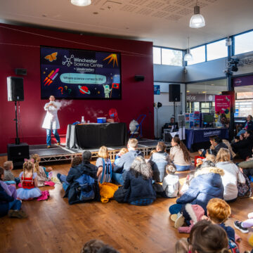 Children and parents are sat on the floor in front of a stage, where a woman is holding a smoking object.