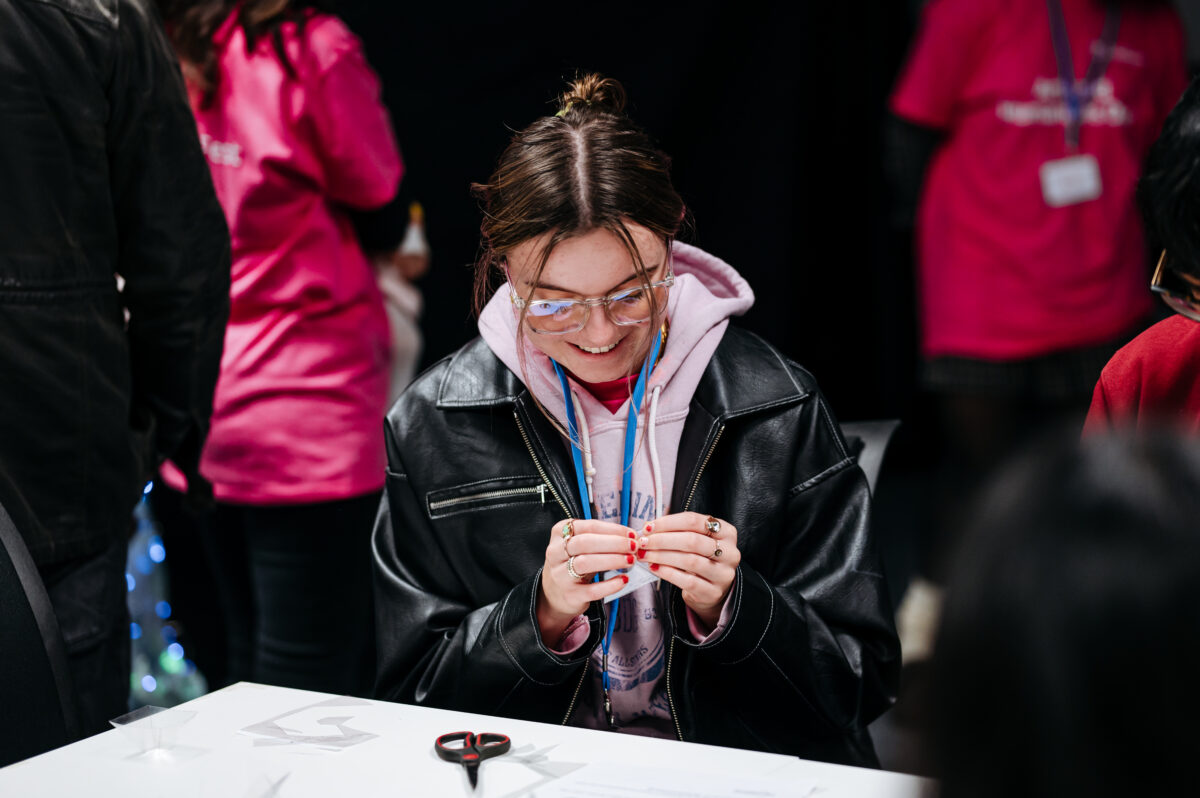 A young woman wearing glasses, a pink hoodie and a black leather jacket looks down at a craft item in her hands