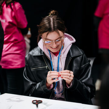 A young woman wearing glasses, a pink hoodie and a black leather jacket looks down at a craft item in her hands