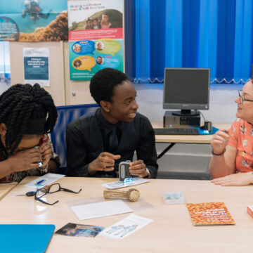 Three people sat at a table talking, the woman on the left is looking down with her hands over her face.