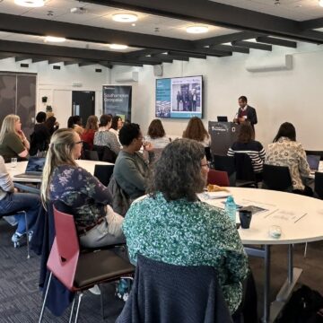 A room full of people sit on white, circular tables looking at a man who is presenting at the front.