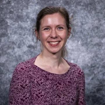 A lady with brown hair in a pony tail and wearing a pink patterned top smiles broadly at the camera