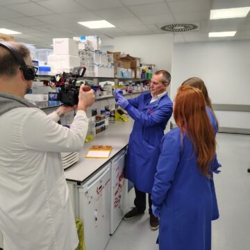 Three people in blue lab coats are stood on the right, whilst a cameraman in a white lab coat records.