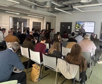 A room full of people sat on light coloured chairs, facing a projector.