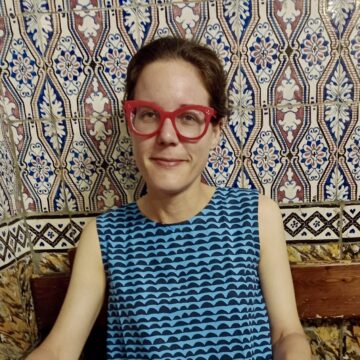 A woman with red glasses and a blue patterned top is smiling.