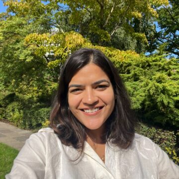 A woman with shoulder-length dark brown hair is smiling in front of some trees.