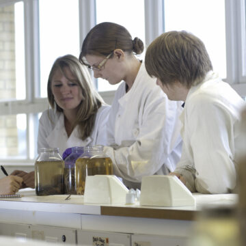 Four people in lab coats are leant over a table with jars of liquid on. The woman to the left is taking notes.