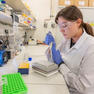 A woman wearing a lab coat and goggles is holding a pipette