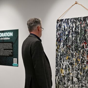 A visitor stands in contemplation of a large painted canvas fabric art work. Further along the wall you can see the sign that welcomes people to the Summer Art Exhibition