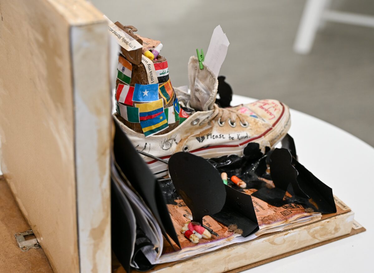 A mixed media art work featuring an old canvas shoe, paper flags, small pegs and mottos arranged in an open book