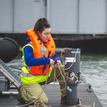 A woman wearing a life jacket is knelt on decking, tying some rope.