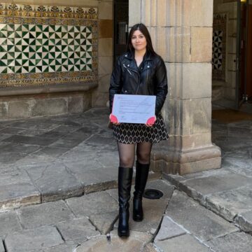 A woman wearing a black leather jacket is stood in a historic building, holding a certificate.