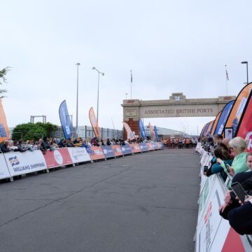 A large group of people are stood behind the horizontal barriers of a running track waiting for the race to begin.