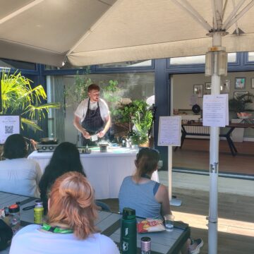 A chef wearing a white shirt and striped blue apron is standing in the corner of a sunny courtyard with a hot plate and cooking ingredients on a table in front of him. A crowd of people are watching him cook